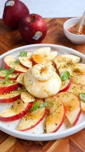 A plate of sliced Cosmic Crisp apples with burrata, hot honey, pistachios and basil, served with toasted bread — whole apples and a honey dipper in the background.