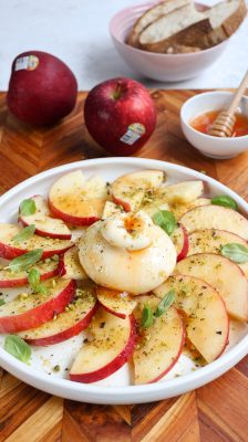 A plate of sliced Cosmic Crisp apples with burrata, hot honey, pistachios and basil, served with toasted bread — whole apples, a honey dipper and sourdough in the background.